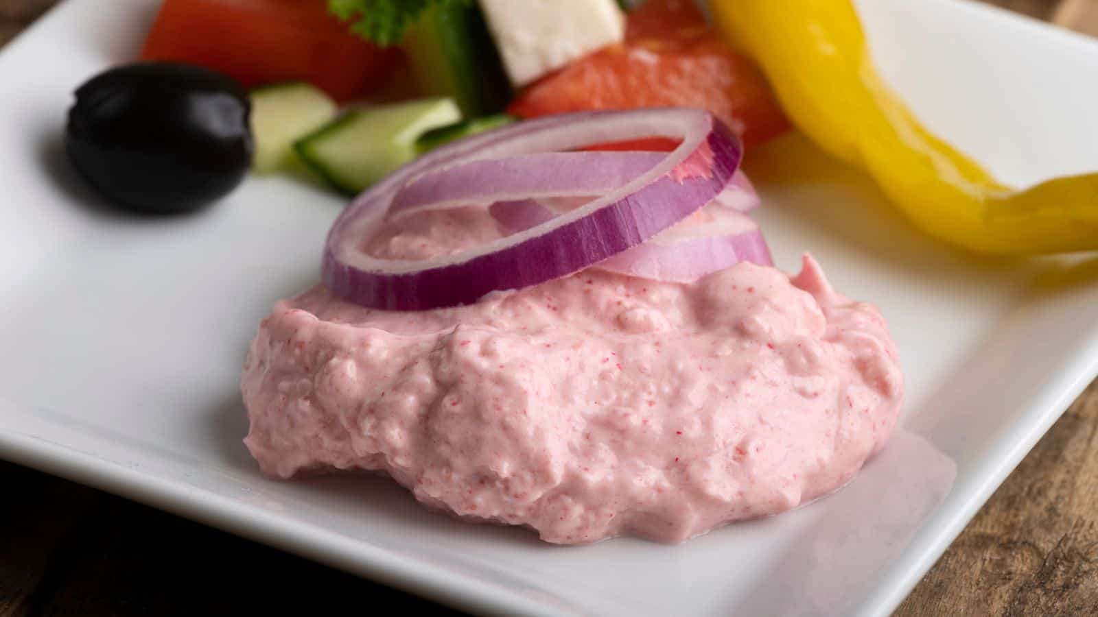 A white plate showcases a pink creamy dip topped with sliced red onions. Nearby, typical of international snacks, are a black olive, sliced cucumber, tomato, and yellow pepper. It's garnished with a parsley sprig and a cube of white cheese for an American twist.