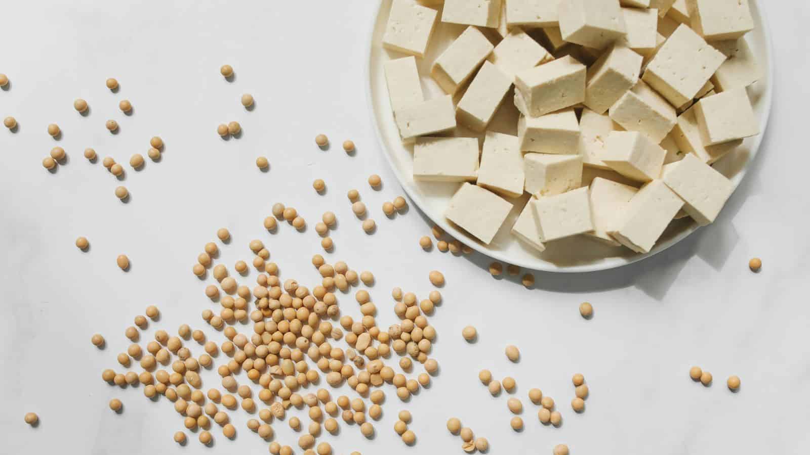 A white plate filled with cubed tofu is placed on a smooth, light surface. Numerous scattered soybeans are spread across the background, creating a contrast between the whole beans and the processed tofu cubes.