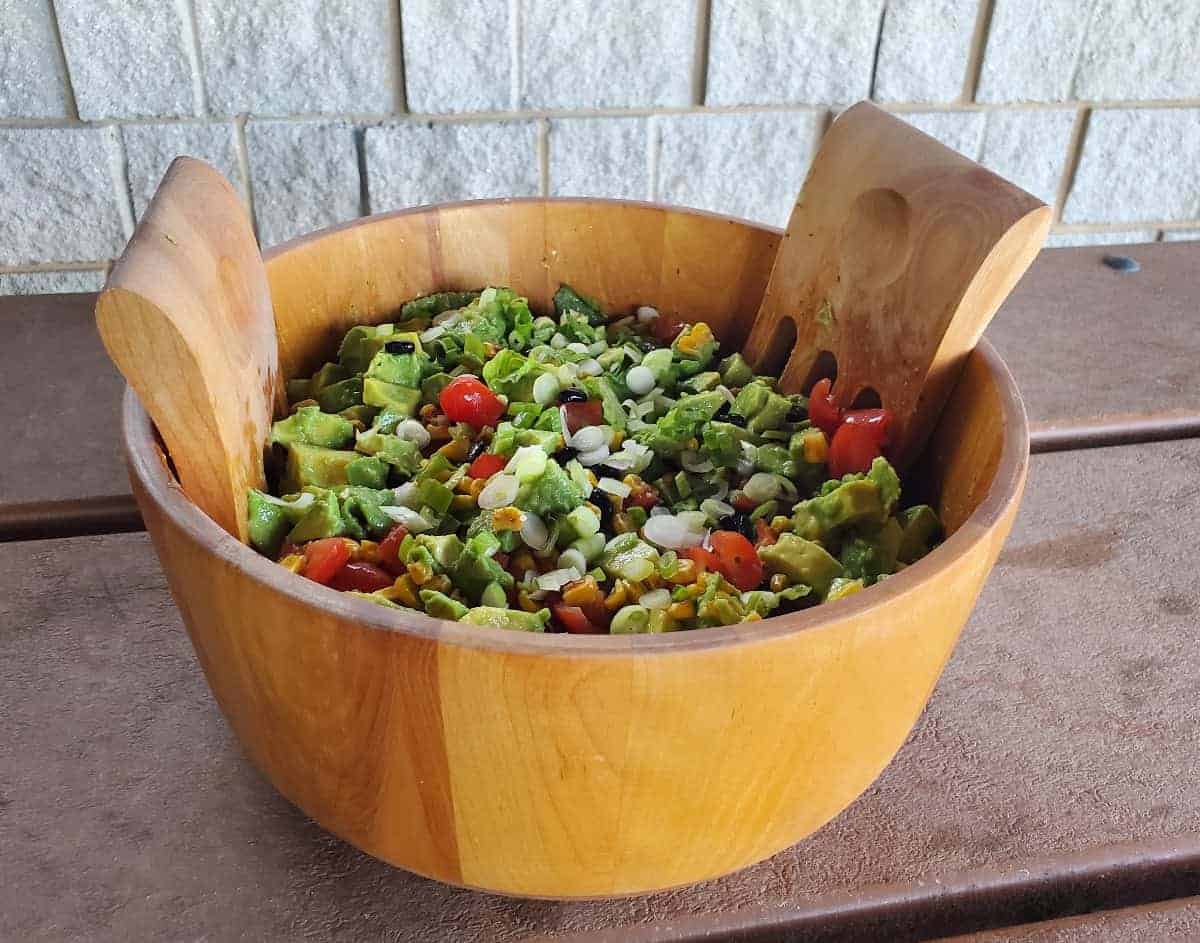 A wooden bowl brimming with a vibrant salad, featuring diced avocado, cherry tomatoes, corn, and green onions. Two wooden salad tossers rest inside. The rustic scene is set against a brick wall backdrop on a wooden surface, making it challenging to overlook such fresh allure.