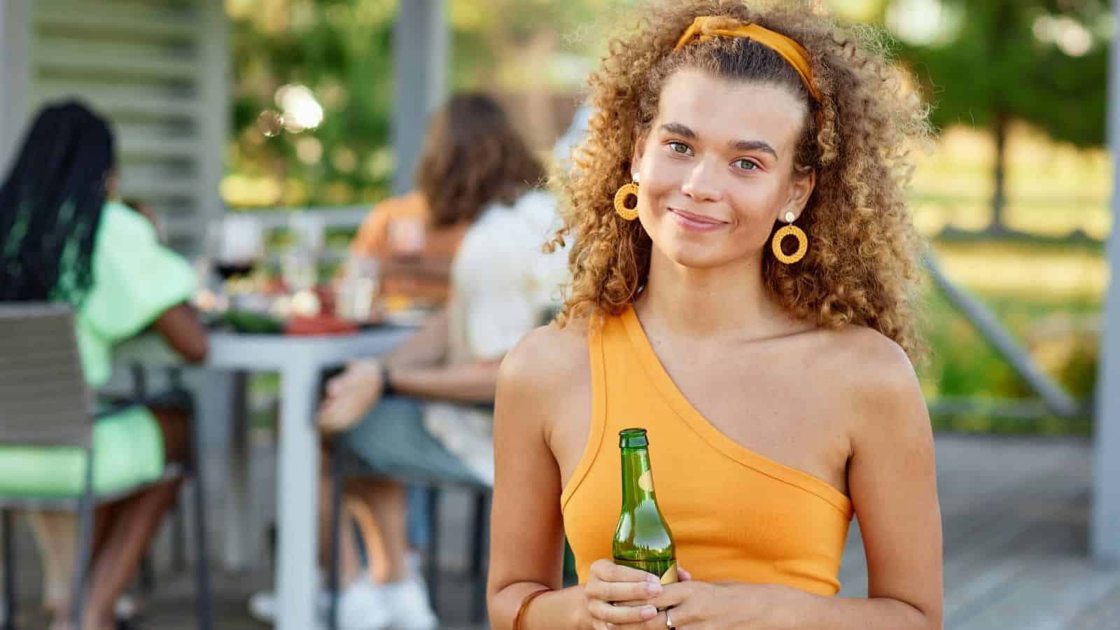 A person with curly hair wearing an orange outfit holds a bottle while standing outdoors. In the background, three people are seated around a table with food and drinks. The setting appears to be a casual gathering on a patio or deck.