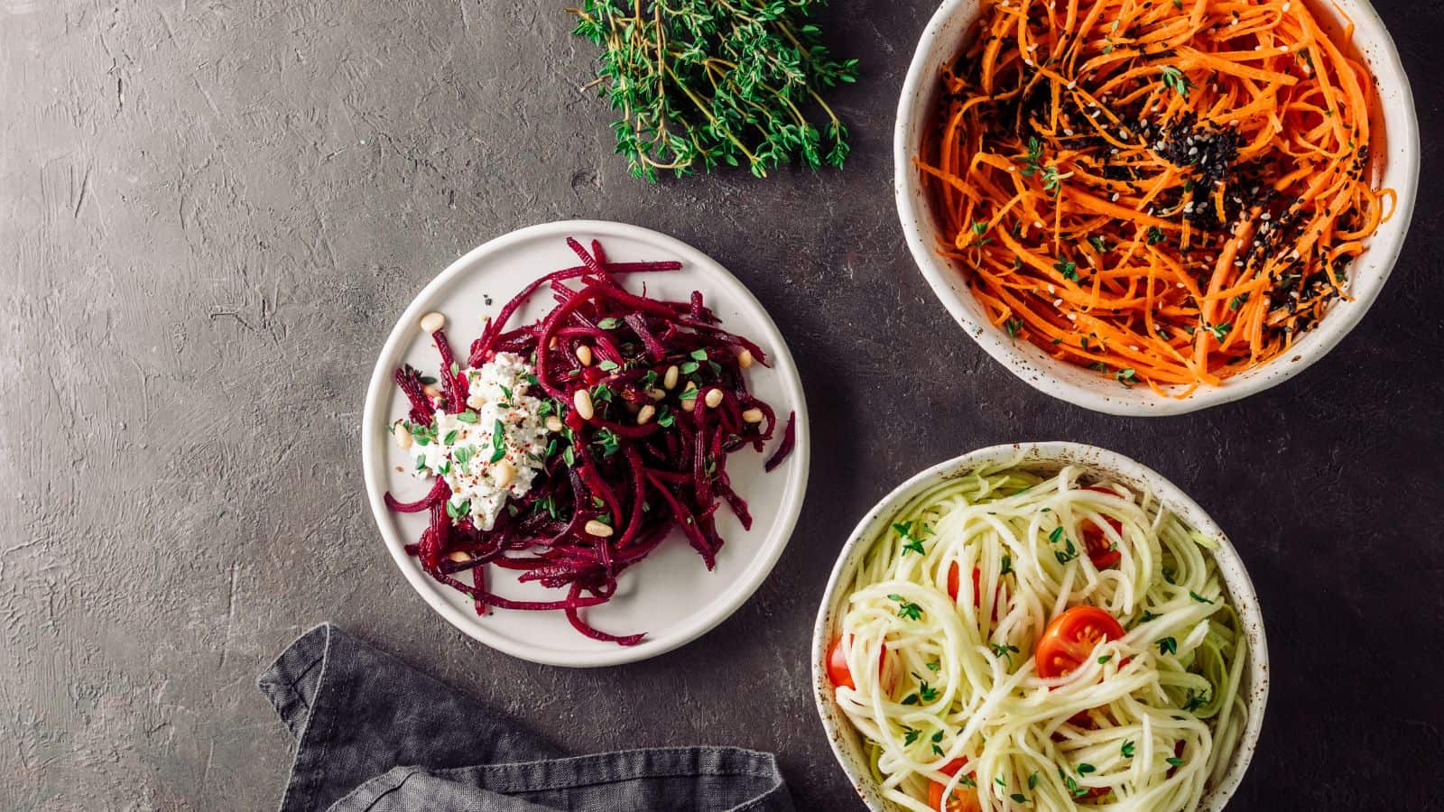 Three bowls of food are displayed on a dark surface. One bowl contains spiralized beets with cheese and seeds, another has spiralized carrots with black seeds, and the third holds spiralized zucchini with cherry tomatoes and herbs. A sprig of thyme is nearby.