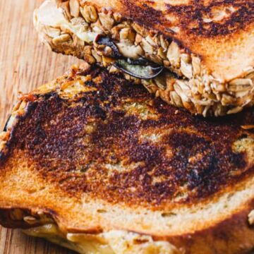 Two grilled cheese sandwiches with crispy, browned tops on a wooden surface. Made with oat-crusted bread and oozing melted cheese, these delights are a brilliant addition to kids' meals. A serrated knife with a light-colored handle sits beside them.