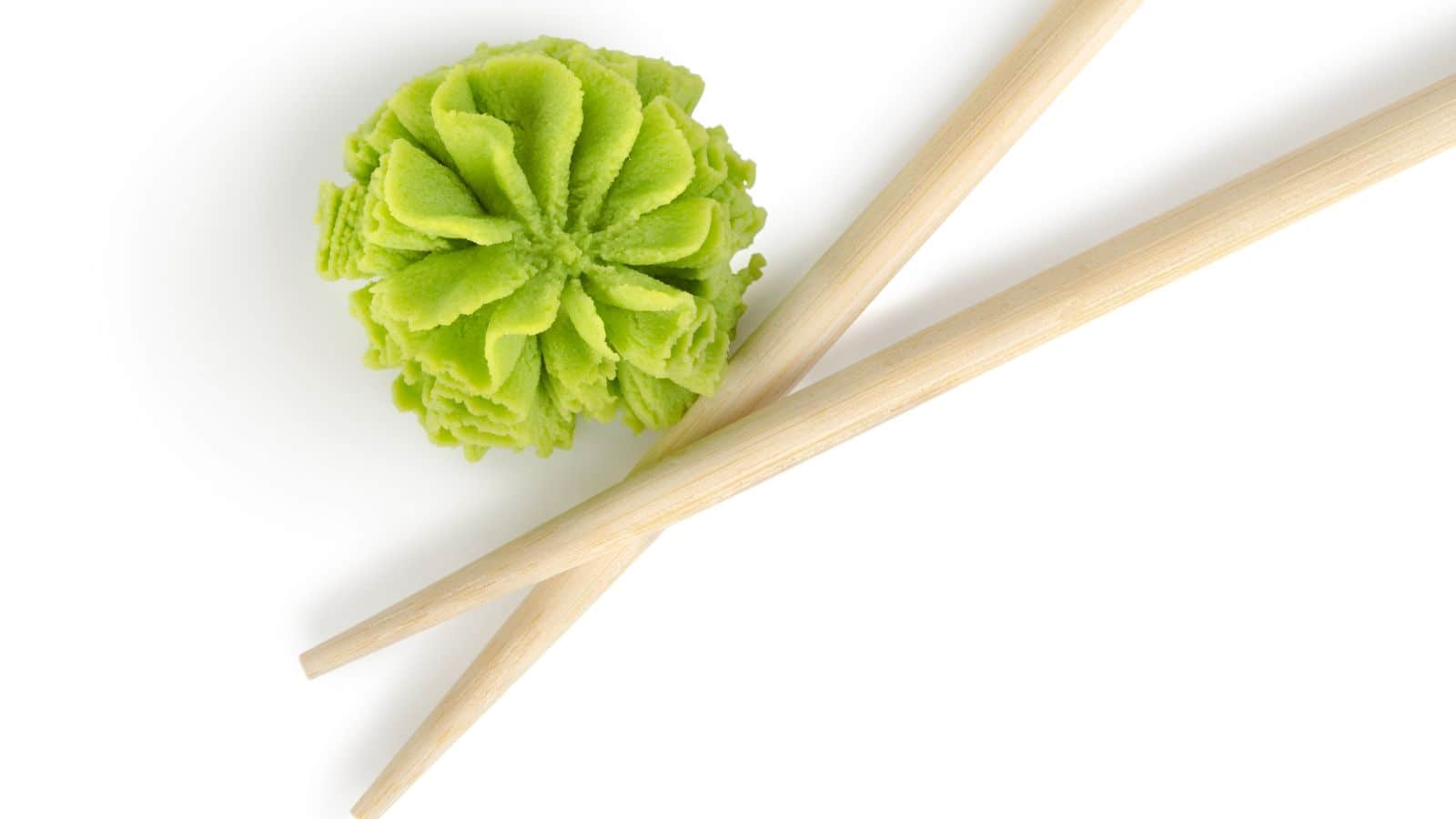 A pair of wooden chopsticks is placed diagonally next to a neatly shaped mound of green wasabi on a white background.