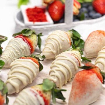 Several strawberries dipped in white chocolate are artfully arranged on a white surface. Some feature a delicate drizzle, while others are dusted with red powder. A small basket with additional strawberries and blueberries sits in the background, perfect for crafting one of 15 no-bake desserts.