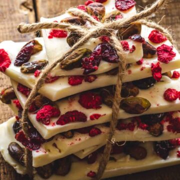 A stack of white chocolate bark pieces tied with string. The bark is topped with dried cranberries, raspberries, and nuts. The stack is placed on a rustic wooden surface.