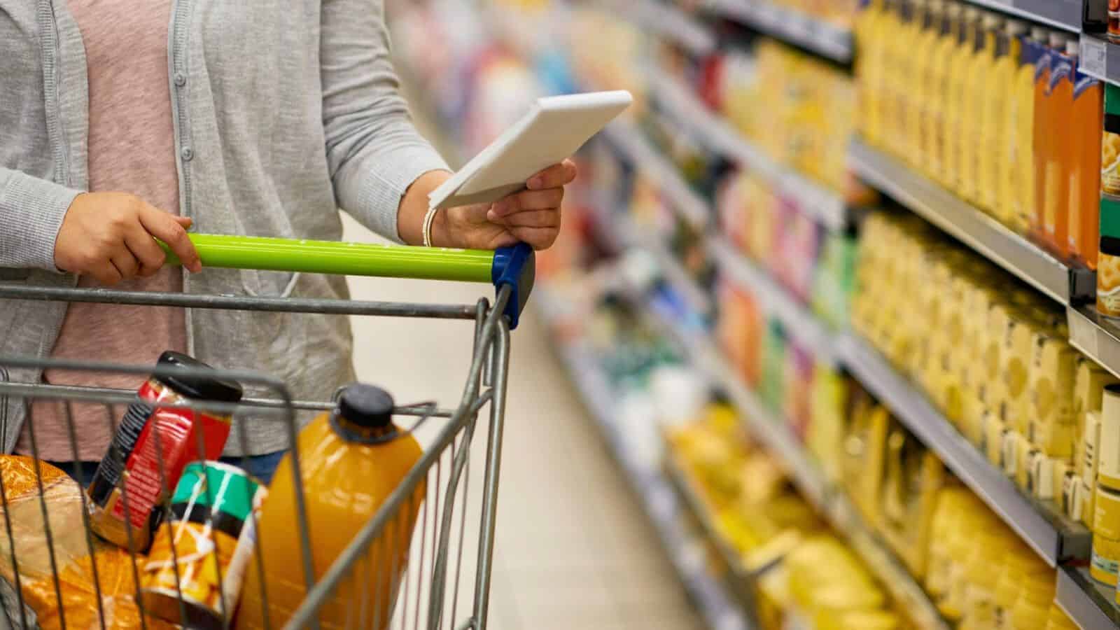 A person pushes a shopping cart filled with groceries in a supermarket aisle. They are holding a white tablet, possibly checking a list or comparing items. The shelves are stocked with various packaged goods and beverages.