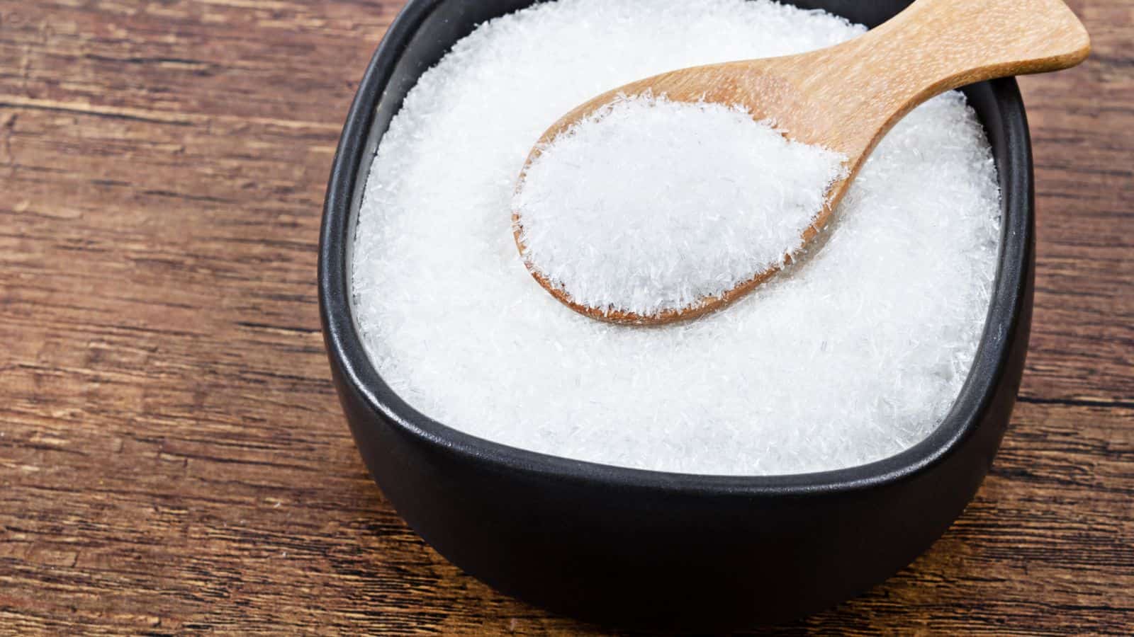 A wooden spoon filled with white, crystalline monosodium glutamate (MSG) rests on the edge of a black bowl containing more of the same. The bowl is placed on a wooden surface, providing a contrast between the white crystals and the dark background.