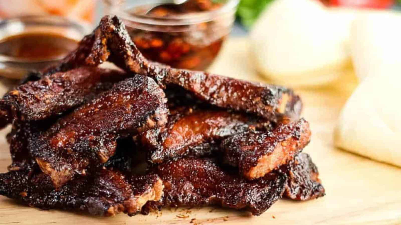 A stack of grilled beef ribs with a shiny, glazed appearance sits on a wooden board—perfect for a Thanksgiving without turkey. Sauces in glass containers are visible in the background, alongside blurred white buns.