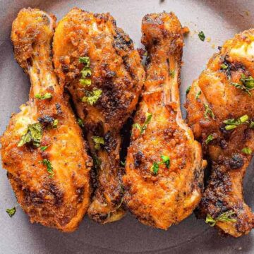 Four seasoned and cooked chicken drumsticks garnished with herbs are placed on a gray plate. The chicken has a golden-brown, crispy appearance.