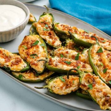 A plate of baked jalapeño poppers, perfect for keto enthusiasts, filled with a creamy cheese mixture and topped with bacon bits. A small bowl of white dipping sauce is on the side. A blue cloth is partially visible in the background, adding charm to these carb-loving delights.