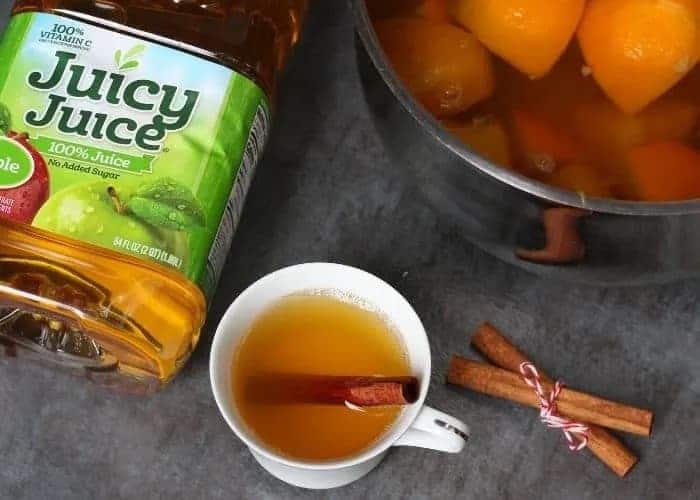 A bottle of Juicy Juice apple juice is placed next to a white teacup filled with a hot drink and a cinnamon stick. Next to the cup, there are three cinnamon sticks tied with string, and an Auto Draft pot containing orange slices and liquid.