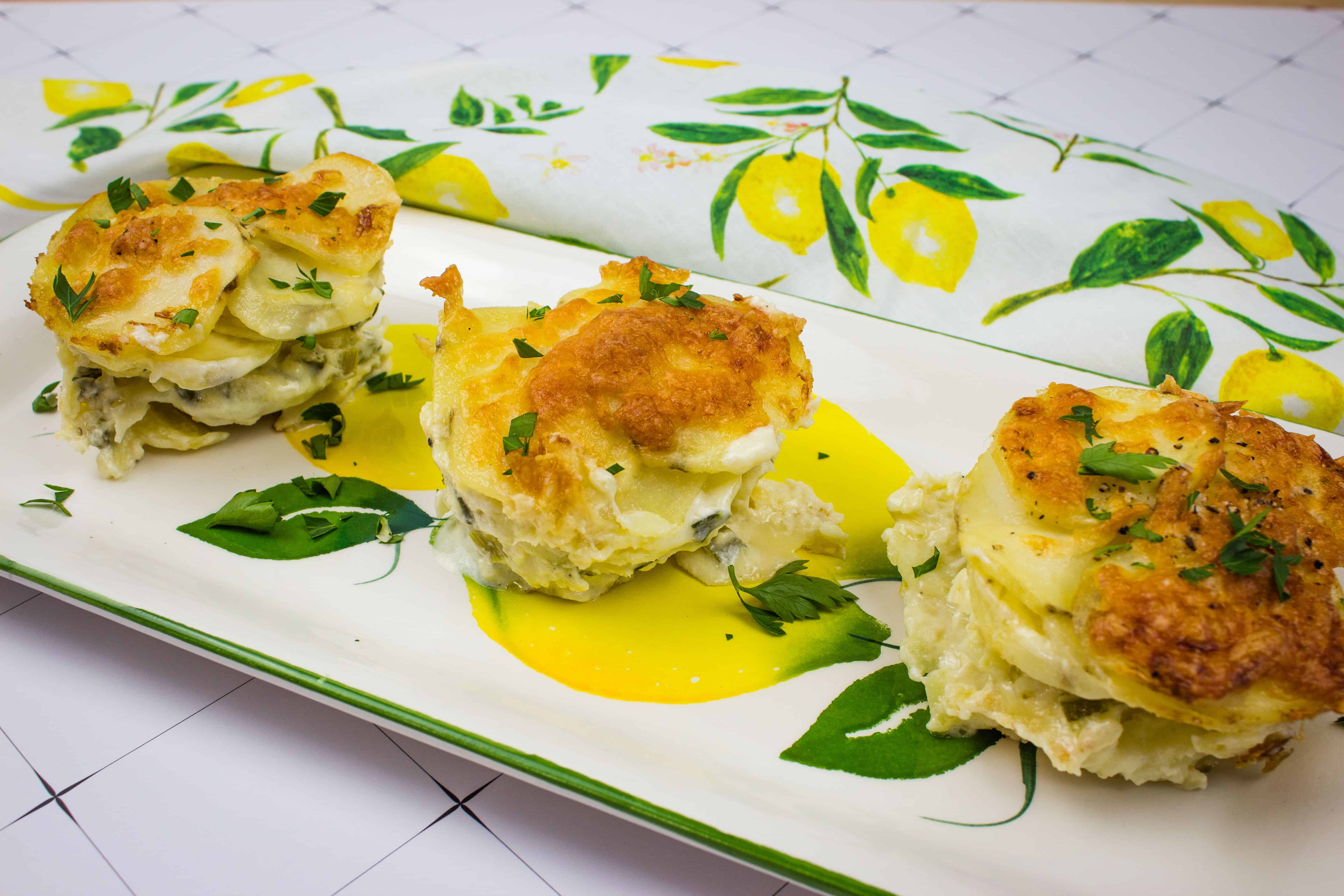 Three servings of potato gratin are artfully arranged on a rectangular plate, their golden-brown crusts garnished with parsley. The plate, resting atop a lemon-patterned cloth, is surrounded by an exquisite yellow sauce against a meticulously tiled surface that seems to be designed with auto draft precision.