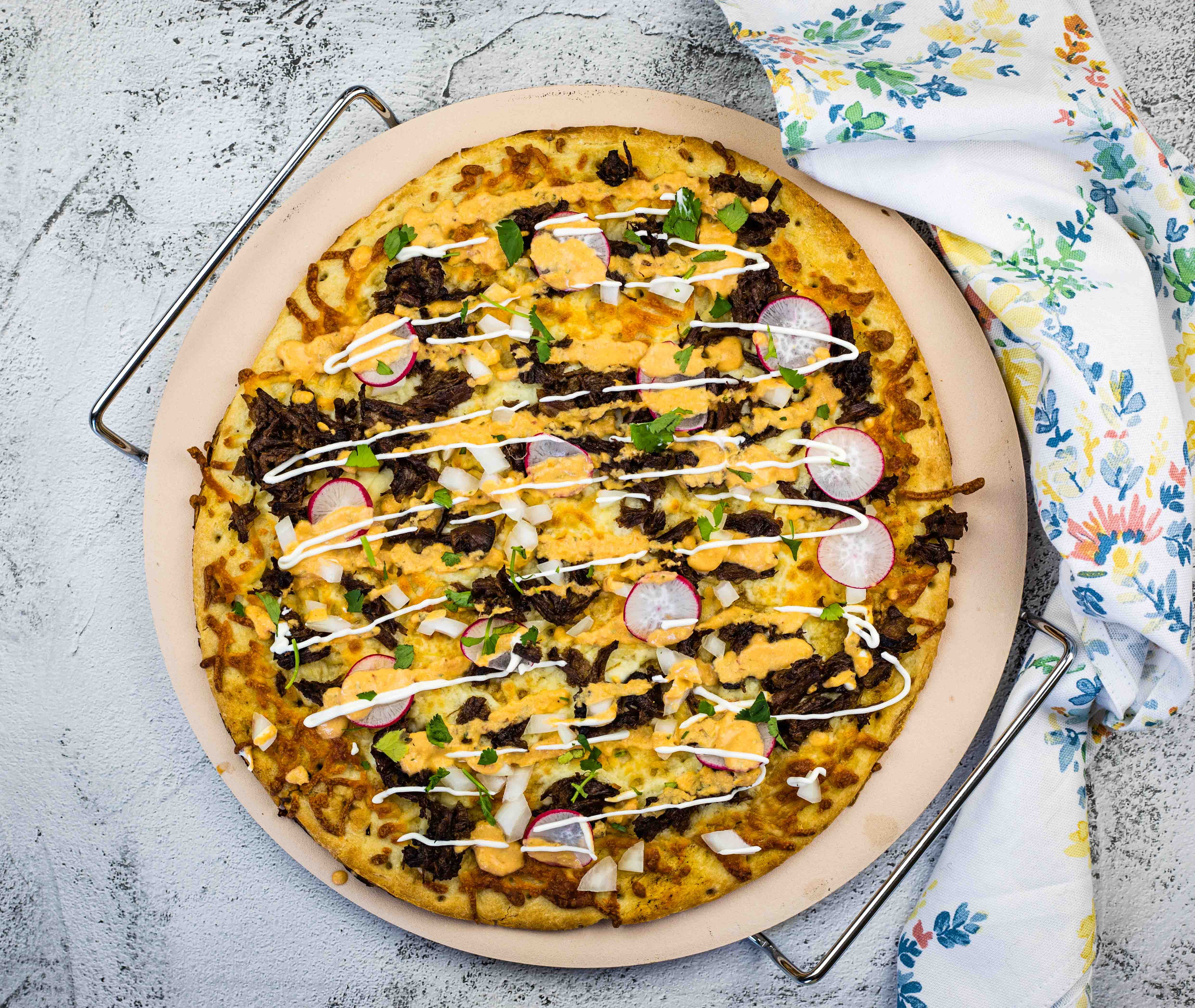 A pizza on a round baking tray topped with beef, radish slices, cilantro, and drizzled with white sauce. A floral napkin is partially visible on the tray's edge. Perfect for exploring new recipes, the backdrop is a textured, light-colored surface.