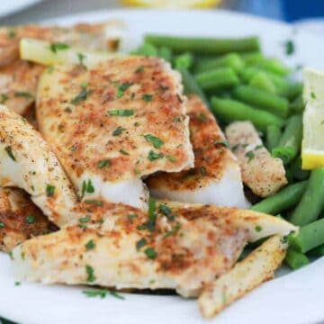 Grilled fish fillets seasoned with spices are served on a plate alongside steamed green beans, making it a perfect keto-friendly dish. A lemon wedge and parsley garnish add a touch of color.