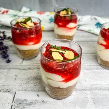 Four small dessert glasses on a white wooden surface, layered with cheesecake filling, a graham cracker base, and topped with raspberry sauce, almond slices, and mint leaves. A bunch of lavender lies nearby on a floral-patterned cloth—a delightful addition to your repertoire of 15 no-bake desserts.