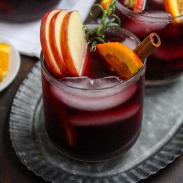 Two glasses of a dark red beverage, garnished with orange slices, apple slices, and cinnamon sticks, are placed on a metal serving tray. This trendy twist on classic favorites leaves you craving more. A white plate with additional orange slices is in the background.