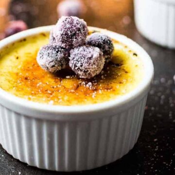 A classic crème brûlée in a white ramekin is topped with sugared cranberries. The custard boasts a caramelized sugar crust and rests on a dark surface adorned with scattered cranberries—perfect for family gatherings. Another ramekin peeks from the background, inviting more delightful moments.