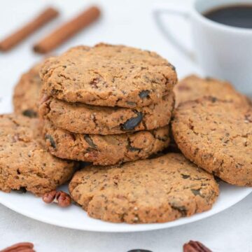 A plate of rustic oatmeal cookies is stacked on a white dish. The cookies contain visible nuts and seeds. In the background, there is a cup of black coffee and a few cinnamon sticks on the table.