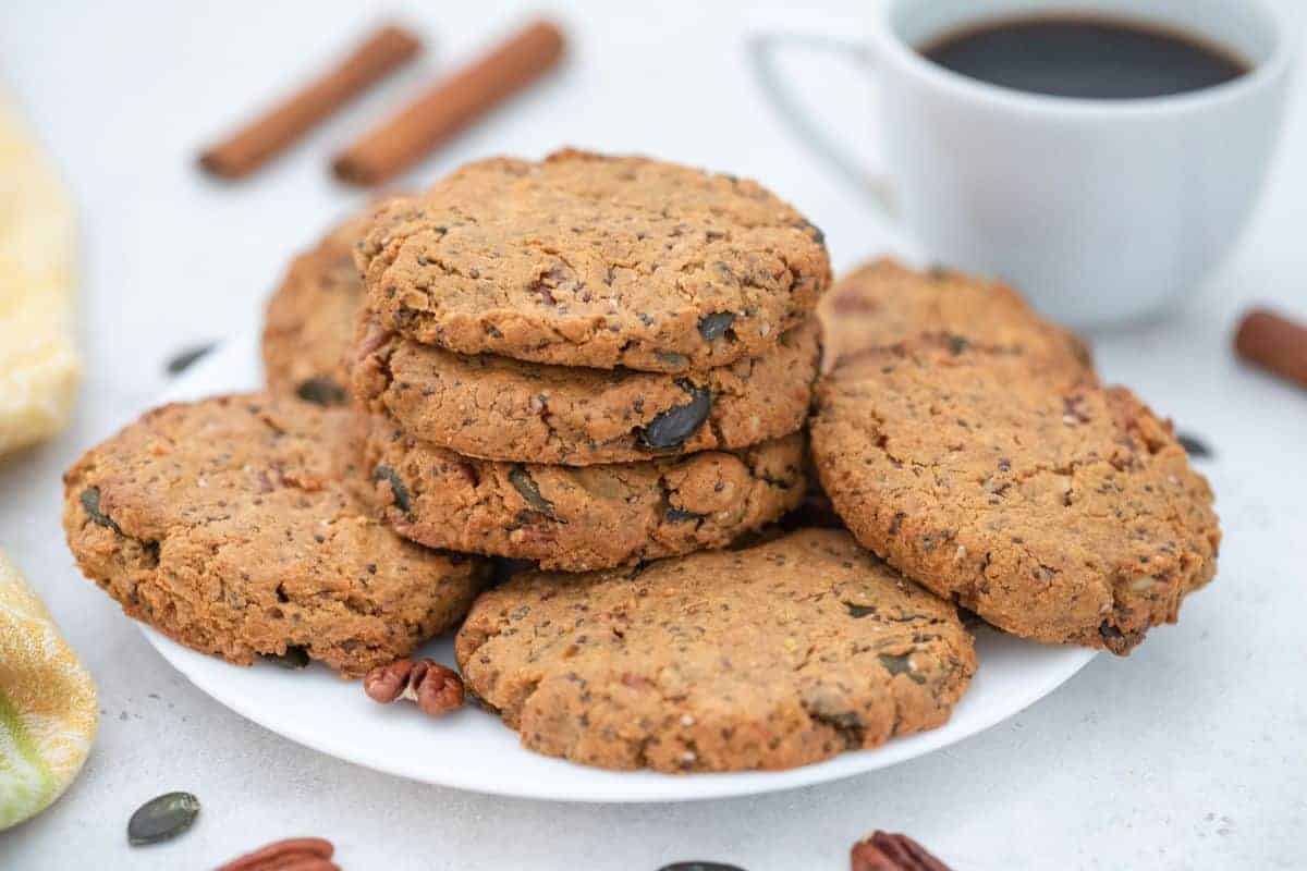 A plate of rustic oatmeal cookies is stacked on a white dish. The cookies contain visible nuts and seeds. In the background, there is a cup of black coffee and a few cinnamon sticks on the table.