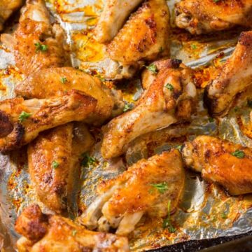 A baking sheet filled with cooked chicken wings, seasoned with spices and garnished with small bits of parsley. The foil beneath the wings is slightly crumpled and shows some browned spots from cooking.