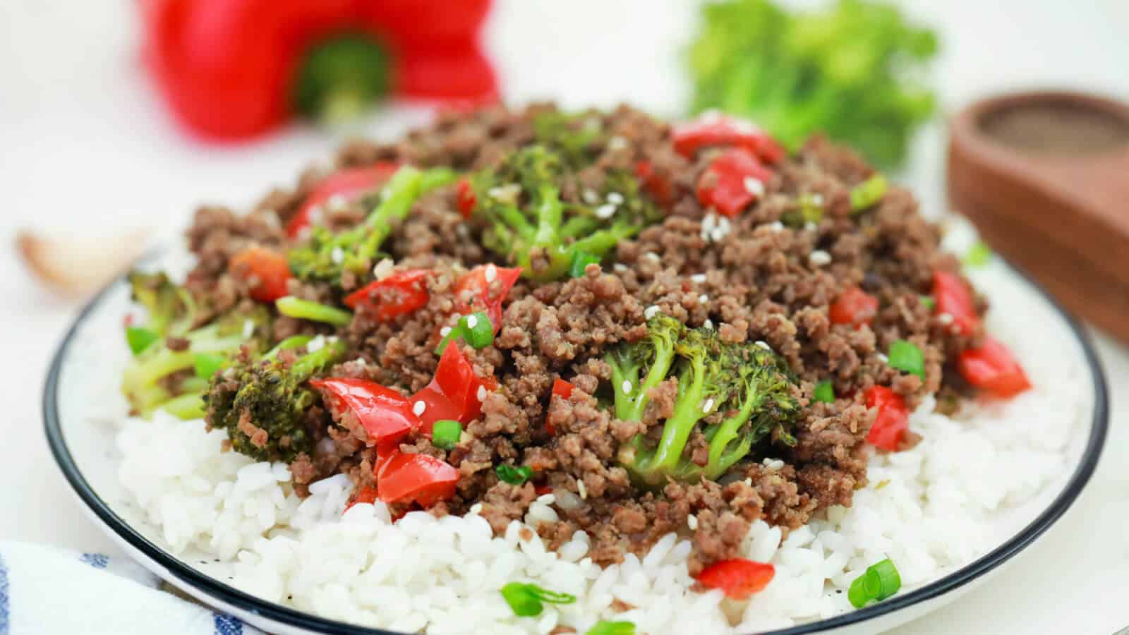 A dish featuring a bed of white rice topped with ground beef, broccoli, and red bell peppers. The meal is garnished with green onions and sesame seeds. A blurred background shows a red bell pepper and broccoli.