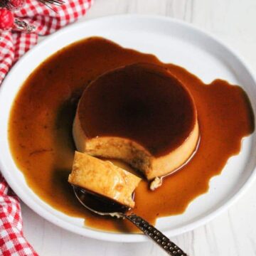 A serving of flan sits on a white plate, enveloped by rich caramel sauce. Beside it, a spoon cradles a delectable piece of the dessert. A red and white checkered cloth adds charm to the scene, suggesting a cozy and inviting atmosphere for enjoying this sweet treat.