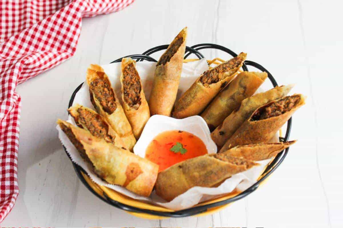 A basket of spring rolls filled with vegetables is arranged around a small square dish of red dipping sauce, garnished with a cilantro leaf. A red and white checkered cloth is partially visible in the upper left corner.