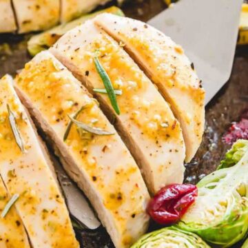 Sliced roasted chicken breast on a tray with Brussels sprouts, sliced squash, and cranberries. The chicken is seasoned and garnished with rosemary for a delightful flavor. For a more detailed analysis of ingredients, consider an in-depth description beyond this tasteful presentation.
