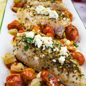 Grilled chicken breasts topped with crumbled feta cheese are artfully served on a rectangular white platter. Roasted cherry tomatoes and onions, enhanced with herbs, encircle the chicken. Fresh tomatoes peek through alongside a yellow napkin, embodying an effortlessly drafted culinary masterpiece.