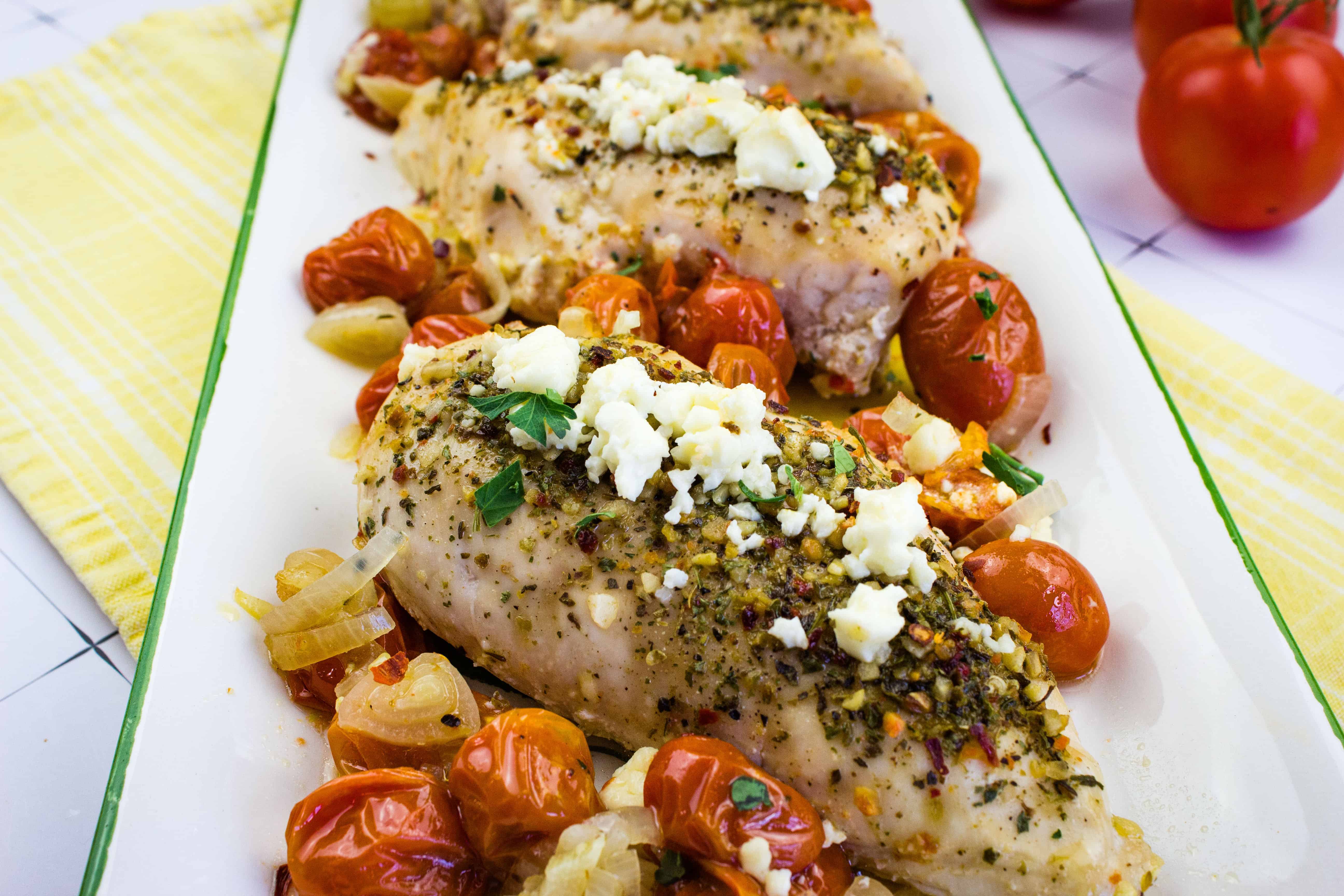 Grilled chicken breasts topped with crumbled feta cheese are artfully served on a rectangular white platter. Roasted cherry tomatoes and onions, enhanced with herbs, encircle the chicken. Fresh tomatoes peek through alongside a yellow napkin, embodying an effortlessly drafted culinary masterpiece.