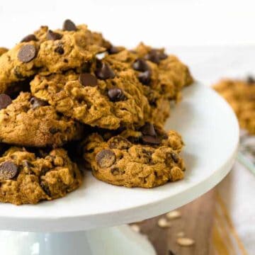 A plate of classic chocolate chip cookies stacked on a white cake stand. The cookies are golden brown with visible chocolate chips, perfect for family gatherings. In the background, more cookies rest on a cooling rack, tempting everyone to skip saving them for later.