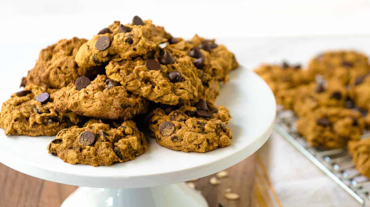 A white cake stand holds a stack of chocolate chip cookies with a golden-brown color and a slightly rough texture. In the background, additional cookies are cooling on an auto draft wire rack atop a wooden surface.
