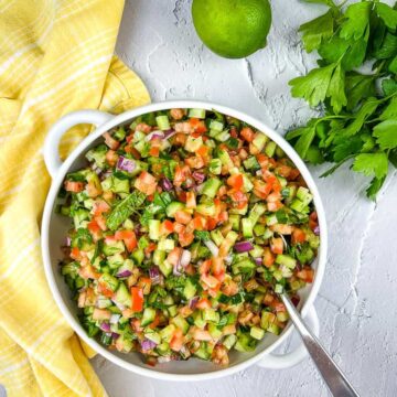 A bowl of finely chopped salad with cucumbers, tomatoes, red onions, and herbs is artfully presented. A silver spoon rests in the bowl, ready to serve. A fresh lime and parsley are beside it on a yellow checkered cloth atop a textured white surface, creating an effortlessly chic auto draft of freshness.