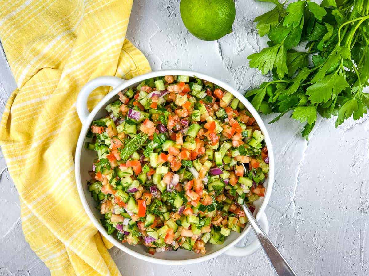 A bowl of finely chopped salad with cucumbers, tomatoes, red onions, and herbs is artfully presented. A silver spoon rests in the bowl, ready to serve. A fresh lime and parsley are beside it on a yellow checkered cloth atop a textured white surface, creating an effortlessly chic auto draft of freshness.