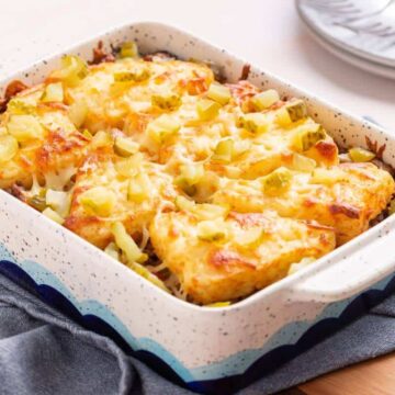 A rectangular casserole dish brimming with baked cheesy tater tot deliciousness, crowned with diced pickles. Resting on a wooden surface layered with a blue cloth, plates and forks await in the background. It's an auto draft of comfort food perfection that promises to satisfy every craving.