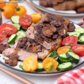 A plate of roasted meat, perfect for keto dishes, is topped with mushrooms and surrounded by sliced cucumbers and halved cherry tomatoes in red and yellow. A wooden pepper container and a red-and-white checkered cloth add charm in the background.