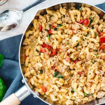 A pan with cooked macaroni mixed with ground meat, spinach, and cherry tomatoes sits on a dark cloth. Fresh basil leaves are to the side, and a jar of spices is partially visible in the corner.