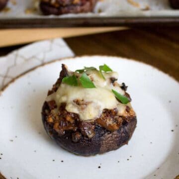 A stuffed mushroom topped with melted cheese and garnished with green herbs sits on a white plate with a brown rim. In the background, more stuffed mushrooms are visible on a baking tray. A napkin lies beside the plate.