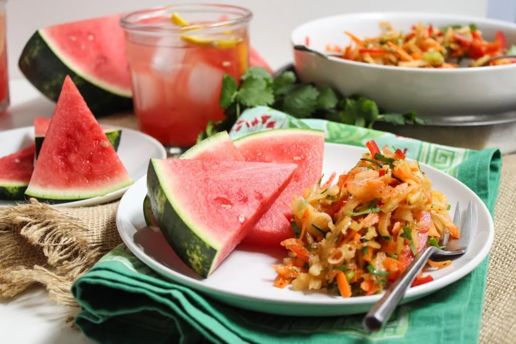 A plate with sliced watermelon and a vibrant carrot and vegetable salad rests on a green napkin. In the backdrop, more watermelon slices accompany a refreshing light pink drink, with fresh cilantro adding a lively touch—an effortless assembly that seems to flow like an auto draft.