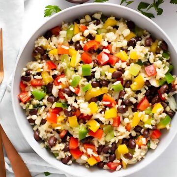 A bowl of colorful salad, reminiscent of favorite holiday festive dishes, features rice, black beans, and vibrant chopped bell peppers in red, yellow, and green. Two copper-colored utensils rest beside the bowl on a white surface. Parsley leaves are scattered around, enhancing its holiday appeal.