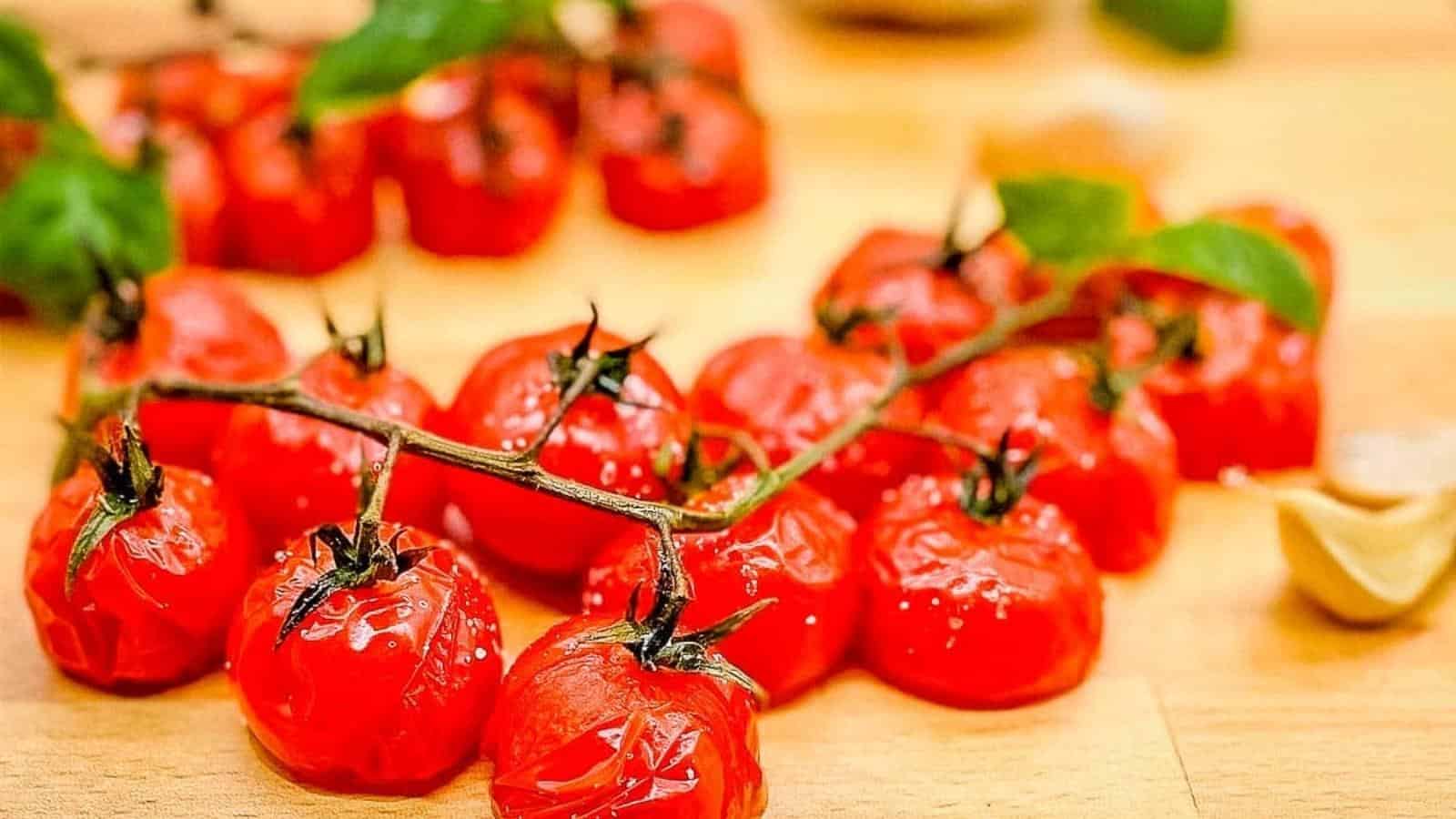 A close-up of vibrant red cherry tomatoes on the vine, perfect for holiday feasts, resting on a light wooden surface. The tomatoes appear fresh with a slight sheen of moisture, making them ideal as side dishes. Some green leaves are attached to the vine.