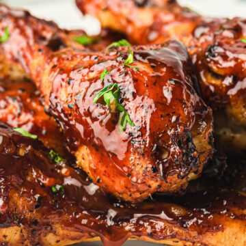 Close-up of grilled chicken drumsticks coated in a glossy barbecue sauce, garnished with small pieces of green herbs. The drumsticks are piled on a white plate.