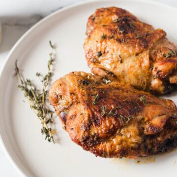 Two cooked chicken thighs with a golden-brown crust seasoned with herbs are placed on a white plate. Sprigs of fresh rosemary are next to the chicken. The background is a white surface with a glimpse of a rustic container.