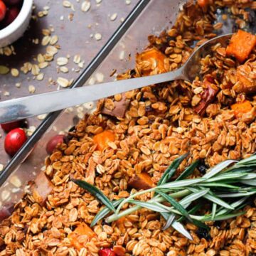 A glass baking dish filled with granola that includes oats and roasted sweet potatoes, garnished with a sprig of rosemary. A spoon rests in the dish. Cranberries and oats are scattered on the wooden surface beside the dish, with a small bowl of cranberries nearby.