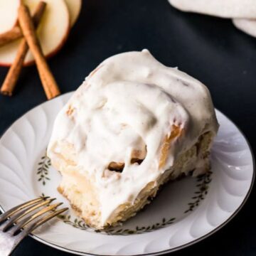 A cinnamon roll with cream cheese frosting sits on a decorative plate. A fork rests beside it. In the background are apple slices and cinnamon sticks on a dark surface.