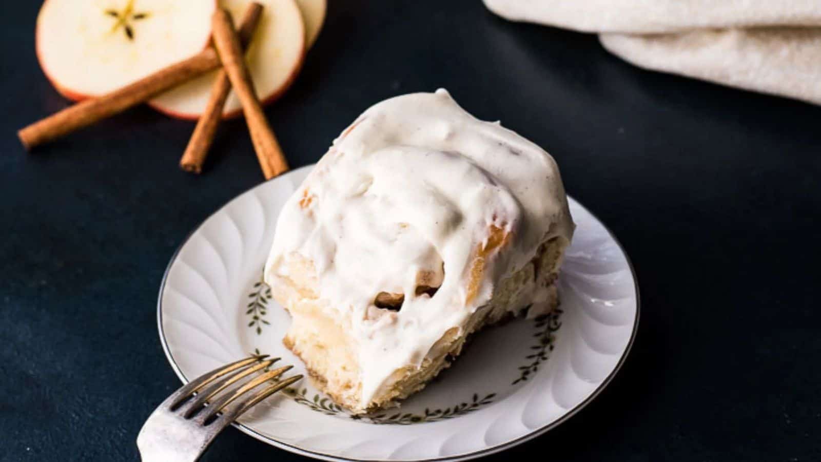 A cinnamon roll with cream cheese frosting sits on a decorative plate. A fork rests beside it. In the background are apple slices and cinnamon sticks on a dark surface.