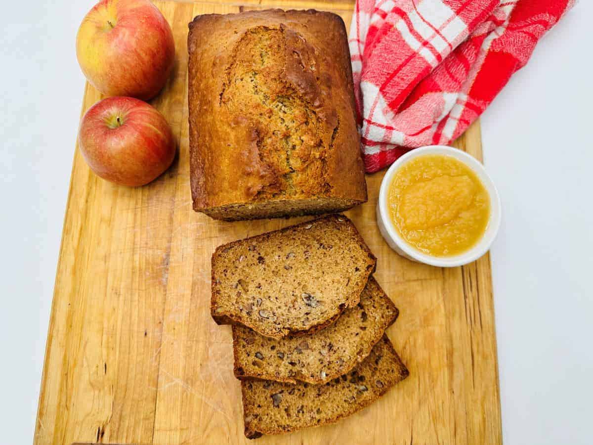 A loaf of holiday banana bread with three slices cut on a wooden board, accompanied by two whole apples, a small bowl of applesauce, and a red and white checkered cloth—perfect side dishes for any festive gathering.