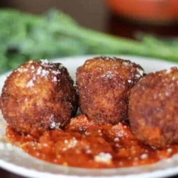 Plate of arancini with marinara and broccolini blurred in background.