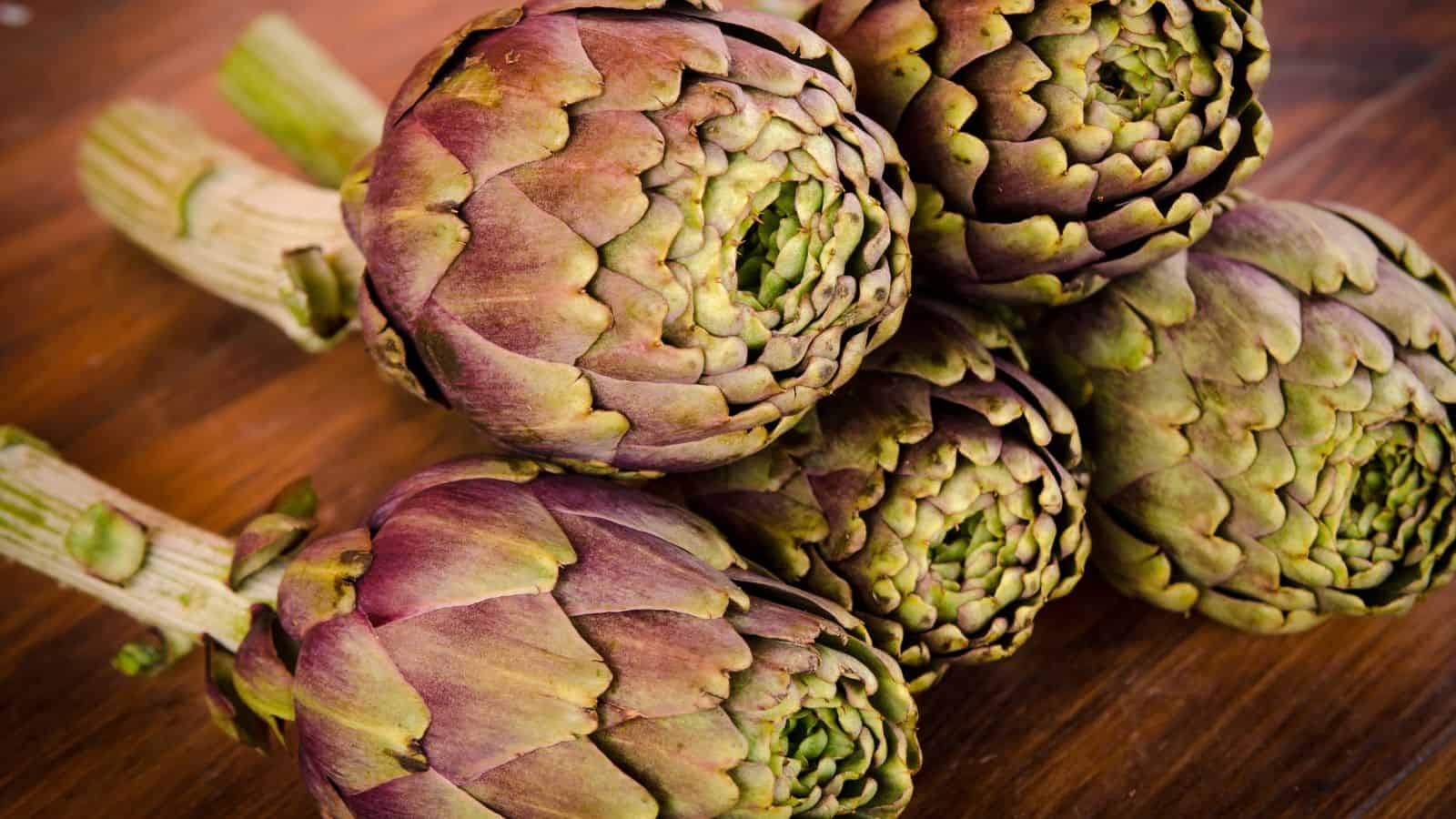 Five fresh artichokes with purplish-green leaves are placed on a wooden surface, showcasing foods rich in fiber. The artichokes are arranged with their stems facing each direction.