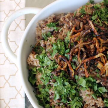 A white dish filled with cooked lentils and rice, topped with chopped cilantro and caramelized onions. A wooden spoon rests on a patterned napkin next to the dish.